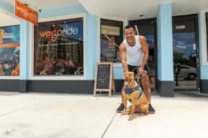 Man smiling with his dog in front of the 'WagPride Pet Boutiques' store entrance in Wilton Manors, FL.